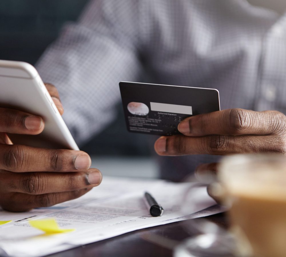 Cropped shot of African-American businessman paying with credit card online making orders via Internet. Successful black male holding plastic card making transaction using mobile banking application